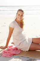 A blonde woman on a beach mat by the sea wearing a white dress