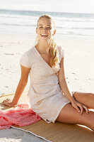 A blonde woman on a beach mat by the sea wearing a white dress