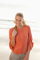 Junge blonde Frau in orangefarbener Bluse und heller Hose am Meer