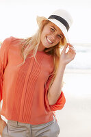 Junge blonde Frau mit Hut in orangefarbener Bluse und heller Hose am Meer