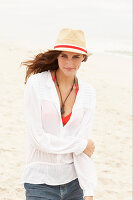 A brunette woman wearing a hat, a red bikini top, a white blouse and denim shorts