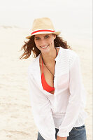 A brunette woman wearing a hat, a red bikini top, a white blouse and denim shorts