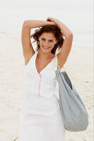 A brunette woman by the sea with a bag wearing a white summer dress