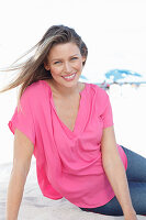 A blonde woman sitting in the sand wearing a pink blouse and jeans