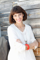 A brunette woman wearing a white long-sleeved blouse standing in front of a wooden wall