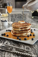 Stack of blueberry buttermilk pancakes with butter adn syrup being poured, paired with mimosa
