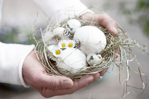 Woman holds small Easter basket made of hay with eggs, daisies and feathers