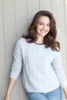 A young brunette woman standing in front of a wooden wall wearing a light knitted jumper and jeans