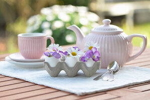 Egg as a vase with horned violets and Marguerite daisies, behind it a tea setting