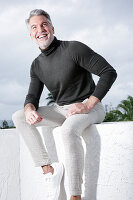 A grey-haired man on a terrace wearing a grey turtleneck jumper and light trousers