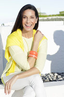 A mature brunette woman wearing a yellow top and white trousers with a jumper over her shoulders