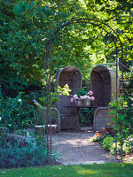 High-backed wicker armchairs on small gravel terrace hidden amongst trees in garden