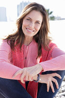 A long-haired woman wearing a pink top and jeans with a jumper over her shoulders