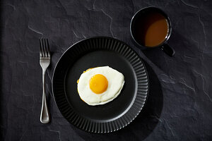 Sunnyside egg with fork and coffee on black surface with black plate and black coffee cup