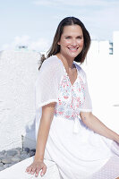 A long-haired woman wearing a white summer dress with colourful embroidery detail