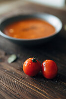 Tomatoes on a wooden surface, with tomato soup in the background
