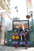 Junge blonde Frau in Daunenjacke, Shirt mit Eulenprint, Karohemd und Jeans vor U-Bahn Schild