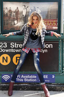 A young blonde woman wearing a down jacket, an owl t-shirt, a checked shirt and jeans standing in front of a Metro sign