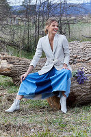 A blonde woman wearing a white blazer and a denim skirt sitting on a fallen tree