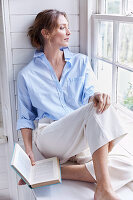 A blonde woman wearing a light-blue shirt-blouse and white trousers sitting on a window sill
