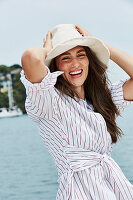 A brunette woman by the sea wearing a hat and a striped shirt-blouse
