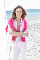 A long-haired woman on the beach wearing a long blouse with a pink jumper over her shoulders