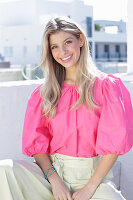 A young blonde woman wearing a pink top with puffed sleeves and white summer trousers