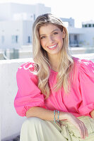 A young blonde woman wearing a pink top with puffed sleeves and white summer trousers