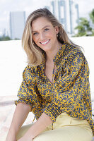 A young blonde woman wearing a black-and-yellow patterned blouse and a pair of light trousers