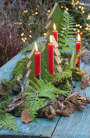 Tischdeko im Wald-Look: Holz-Tannenbäumchen, Zweige mit Moos, Farnblätter, Herbstlaub und 4 rote Kerzen, im Hintergrund Lichterkette