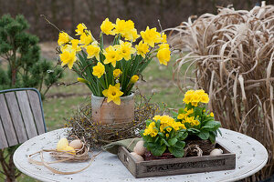 Bouquet of daffodils in a wreath of twigs, primroses in baskets on a tray, and Easter eggs on the patio table