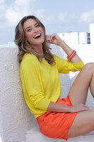 A dark-blonde woman wearing a yellow blouse and orange shorts