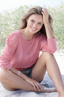 A young, long-haired woman wearing a pink blouse and shorts sitting in the sand