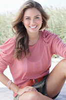 A young, long-haired woman wearing a pink blouse and shorts sitting in the sand