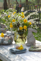 Ostertischdeko im Garten: Strauß aus Blütenzweigen und Löwenzahn, Osterhase, Korb mit Ostereiern und Teller mit Serviette