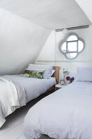 Two single beds with light-colored bed linen in sloped ceiling room with porthole window