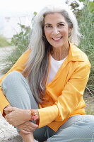 A mature woman with grey hair wearing an orange blouse and trousers sitting on a beach