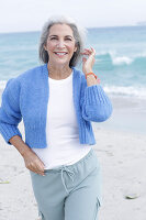 Mature woman with grey hair in white t-shirt, blue cardigan and trousers on the beach