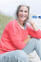 A mature woman with grey hair wearing a salmon-pink jumper and trousers on a beach