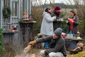 Man puts log in fire pit, in the background women with mulled wine