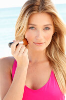 Young blonde woman in pink dress applying makeup with brush