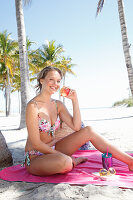 Junge blonde Frau im Bikini sitzt auf pinkfarbener Decke unter einer Palme am Strand