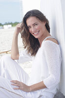 A long-haired woman on the beach wearing a white jumper and white trousers