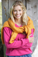 Junge blonde Frau in pinkfarbenem Pullover, mit gelbem Strickpulli über den Schultern vor Bretterwand