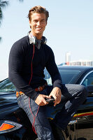 A young man wearing a jumper standing next to a car with headphones and a telephone