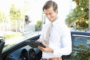 A young businessman holding a tablet computer standing next to a car