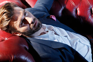 Young man with a beard in a jacket and white shirt lying on a leather sofa