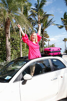 Young blond woman in a pink sundress stands in the skylight opening of a car