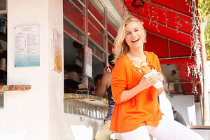Junge blonde Frau in orangefarbener Bluse isst Sandwich auf der Straße