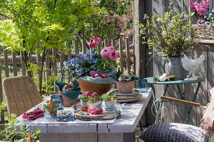Easter eggs with cress in glasses, bread basket and flower bowl with ranunculus and forget-me-not, cutlery, crockery and wooden disc on garden table